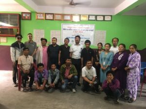 The image shows a group of people posing for a group photo indoors, after the event. They are standing and sitting in two rows in front of a green wall with certificates and a banner in the background. The banner contains text in Nepali and English, mentioning the name of the event, partners and other details. The group consists of both men and women of various ages, and everyone appears to be smiling or in a positive mood.