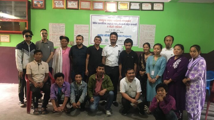 The image shows a group of people posing for a group photo indoors, after the event. They are standing and sitting in two rows in front of a green wall with certificates and a banner in the background. The banner contains text in Nepali and English, mentioning the name of the event, partners and other details. The group consists of both men and women of various ages, and everyone appears to be smiling or in a positive mood.