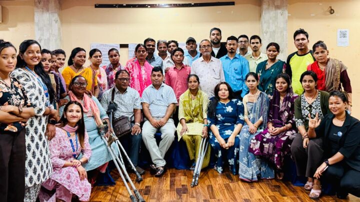 This is a group photo featuring a diverse mix of men and women, some of whom are persons with disabilities. They are gathered indoors, standing and seated together, smiling at the camera. The setting is the orientation session conducted at Janankpur.