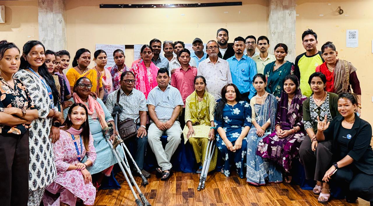 This is a group photo featuring a diverse mix of men and women, some of whom are persons with disabilities. They are gathered indoors, standing and seated together, smiling at the camera. The setting is the orientation session conducted at Janankpur.