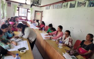 This image captures a session, involving women with disabilities seated around a U-shaped table arrangement. The participants are actively engaged in discussions and note-taking. Some participants are writing on large sheets of paper, indicating group activities or brainstorming tasks. The presence of notebooks, pens, and bottled water suggests that the session is designed to be interactive. A ceiling-mounted projector and visual displays on the walls hint at the use of multimedia or visual aids during the session. The overall atmosphere is one of active participation, learning, and empowerment within a supportive community setting.