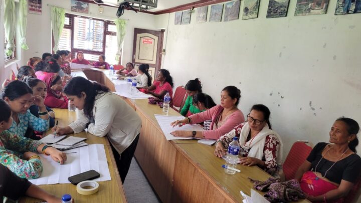 This image captures a session, involving women with disabilities seated around a U-shaped table arrangement. The participants are actively engaged in discussions and note-taking. Some participants are writing on large sheets of paper, indicating group activities or brainstorming tasks. The presence of notebooks, pens, and bottled water suggests that the session is designed to be interactive. A ceiling-mounted projector and visual displays on the walls hint at the use of multimedia or visual aids during the session. The overall atmosphere is one of active participation, learning, and empowerment within a supportive community setting.