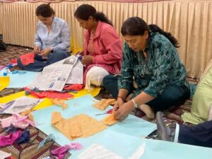 There are three women sitting next to each other on the training hall floor. There are chart papers in front of them. The women are cutting different pieces of clothes and making different figures they attach into the chart paper.