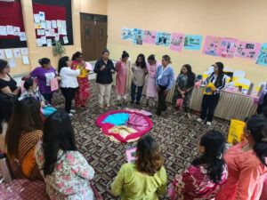 The participants are standing in a circle inside a room. There are colorful clothes kept in a circular shape. A woman in black top is sharing her story with the group. The walls have different drawings and postcards made by the participants.