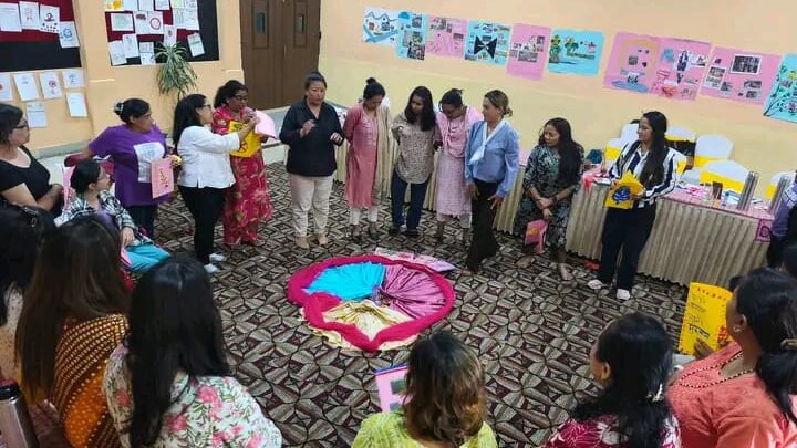 The participants are standing in a circle inside a room. There are colorful clothes kept in a circular shape. A woman in black top is sharing her story with the group. The walls have different drawings and postcards made by the participants.