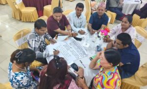 A group of people are sitting on a round table in a circle. They have spread chartpapers in front of them. The participants are discussing and writing on the chartpaper.