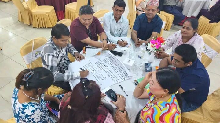 A group of people are sitting on a round table in a circle. They have spread chartpapers in front of them. The participants are discussing and writing on the chartpaper.