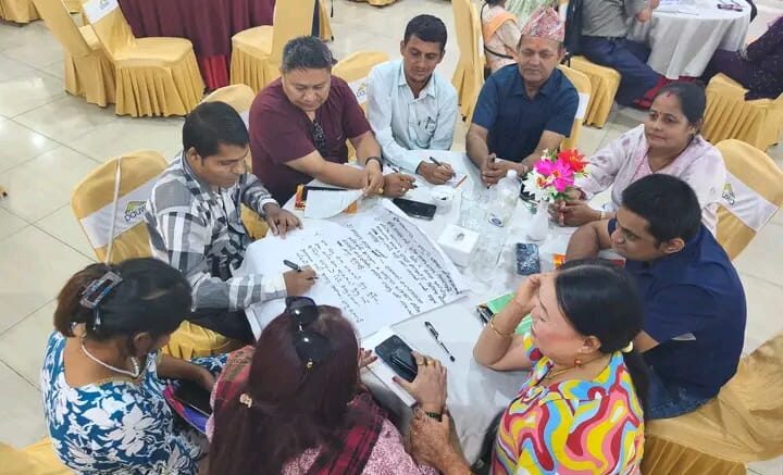 A group of people are sitting on a round table in a circle. They have spread chartpapers in front of them. The participants are discussing and writing on the chartpaper.