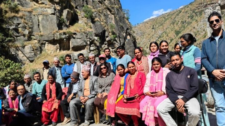 A large group of people, including men and women, are gathered for a group photo outdoors after the session is completed. Some are seated in the front row while others stand behind them, with rocky cliffs and hills of Kalikot visible in the background under a clear blue sky.