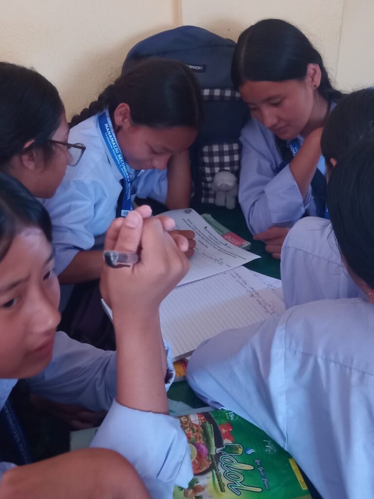 The adolescent girls are discussing. One of them have paper laid in front of her and she is writing something. Other girls are sitting around her.