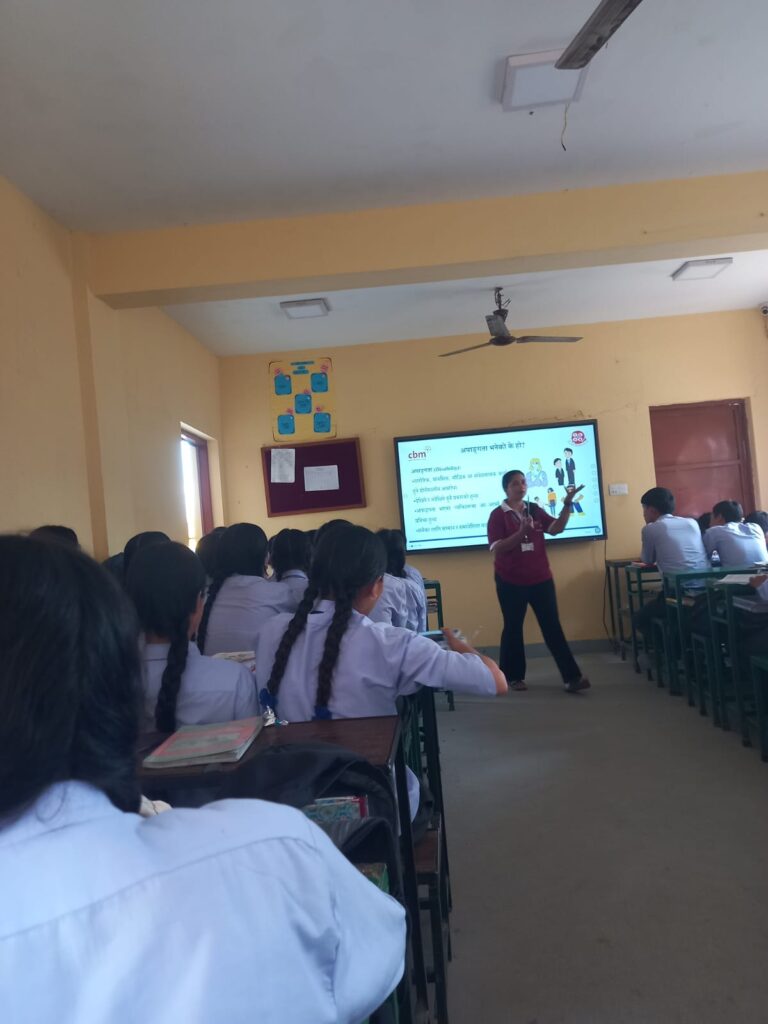 The facilitator is facing towards the camera at a distance. The presentation is on a big screen behind her. The students are facing towards the screen, listening to the facilitator.