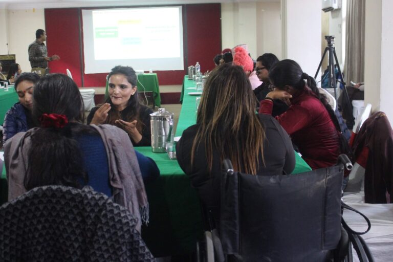 The picture is in a room-setting. There is a white screen at the back and there is presentation in the screen. Two sign language interpreters are facing the camera and the participants are discussing. The presenter is reading the slide.