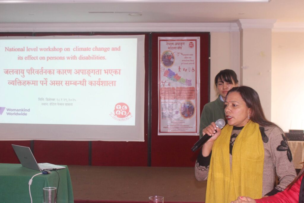 Ms Meena Paudel is speaking on mic during an event. The banner behind her shows the program is related to disability and climate change.