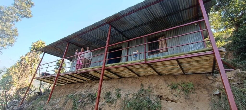 Kamala Phuyal and Pramila Shrestha are posing towards camera. The house is made up of tin sheets and is supported by iron frames.