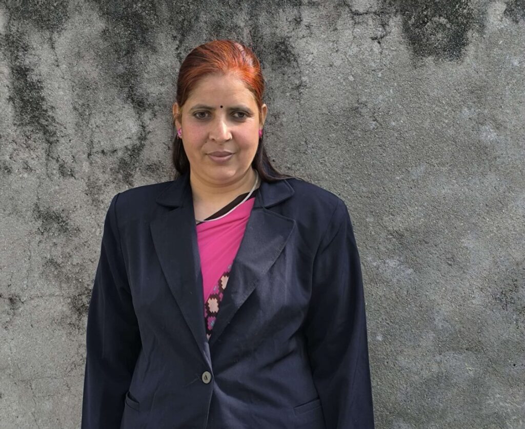 Devi is posing for the photo. She is wearing a black blazer and pink sari, which is uniform of teacher.