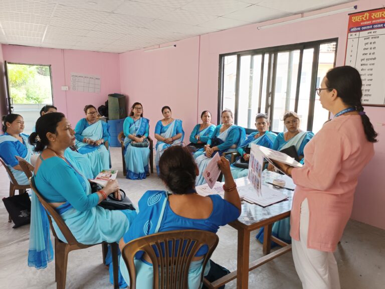 The FCHVs are sitting in a round setting. Ms. Debina is standing in front and is explaining about GBV and disability using flipchart.