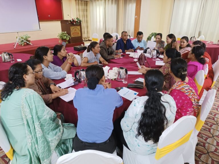 The participants are sitting in a circle on table. They have notebooks in front of them and are involved in discussion.